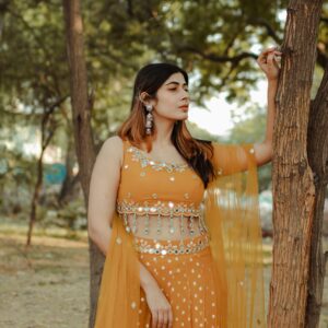 A young Indian woman in an elegant orange gown poses gracefully outdoors.